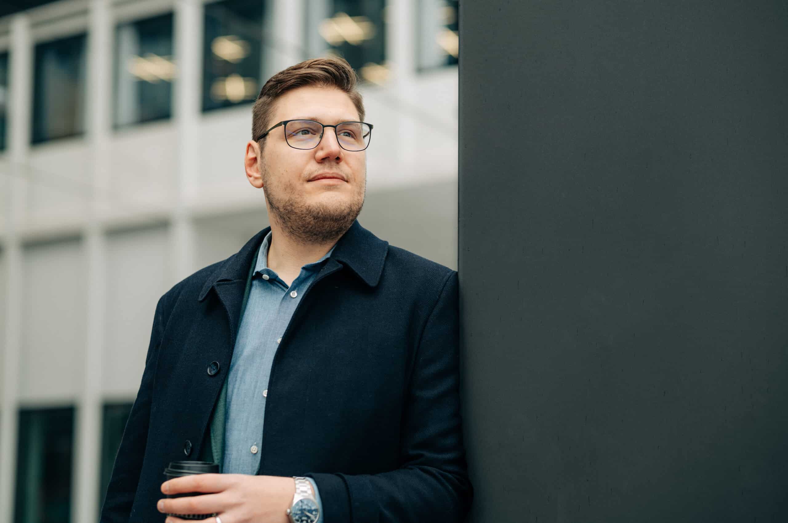 Pascal Turin lehnt an eine Wand. Er trägt eine Brille und einen Mantel, darunter ein Hemd. In der Hand hält er einen Coffee-to-Go-Becher. Zu sehen ist auch seine Uhr der Marke Oris am Arm.