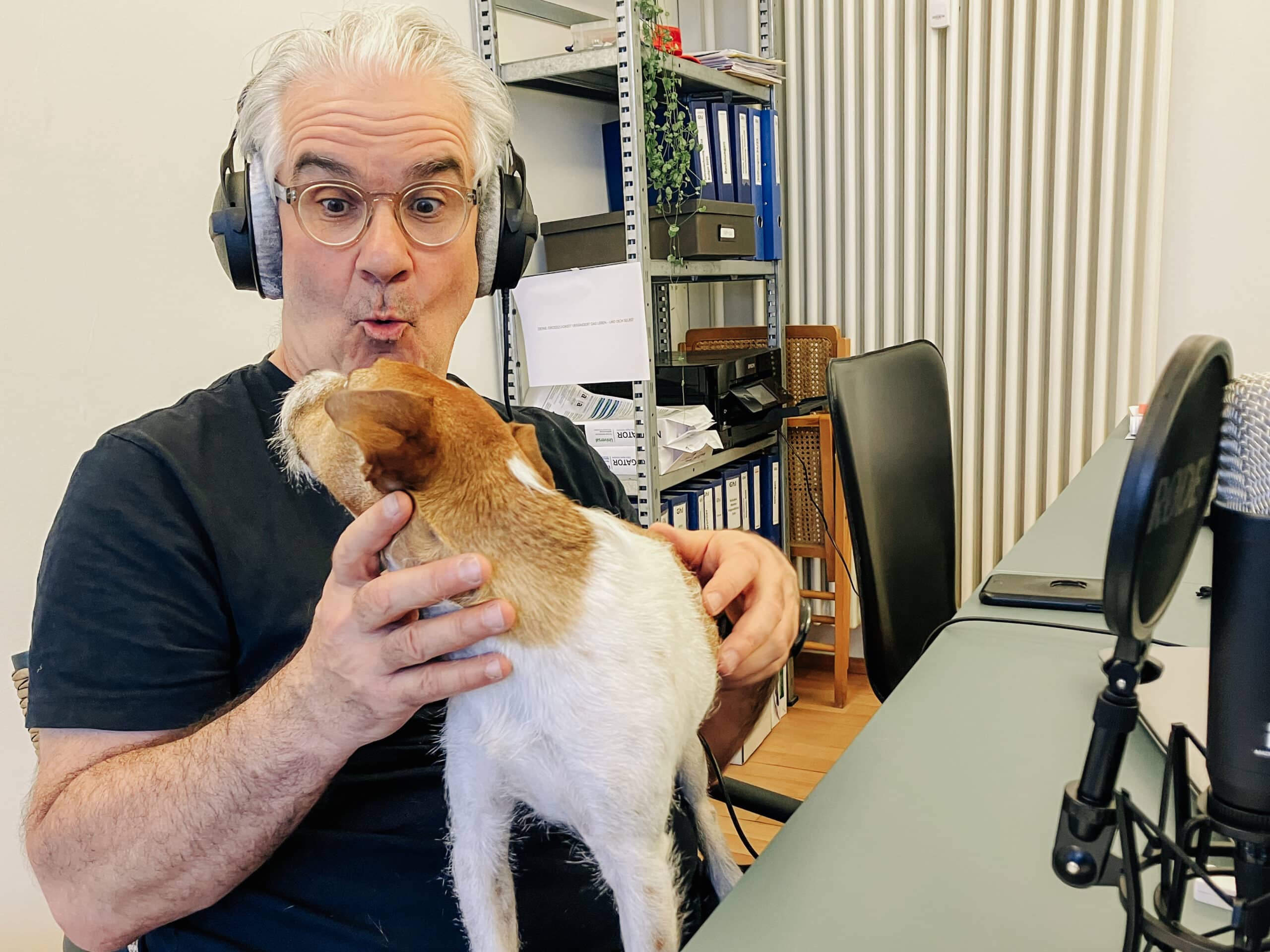 Lorenz Steinmann sitzt vor einem Podcast-Mikrofon und trägt Kopfhörer. Auf seinem Schoss steht der Parson Russell Terrier Waldo.