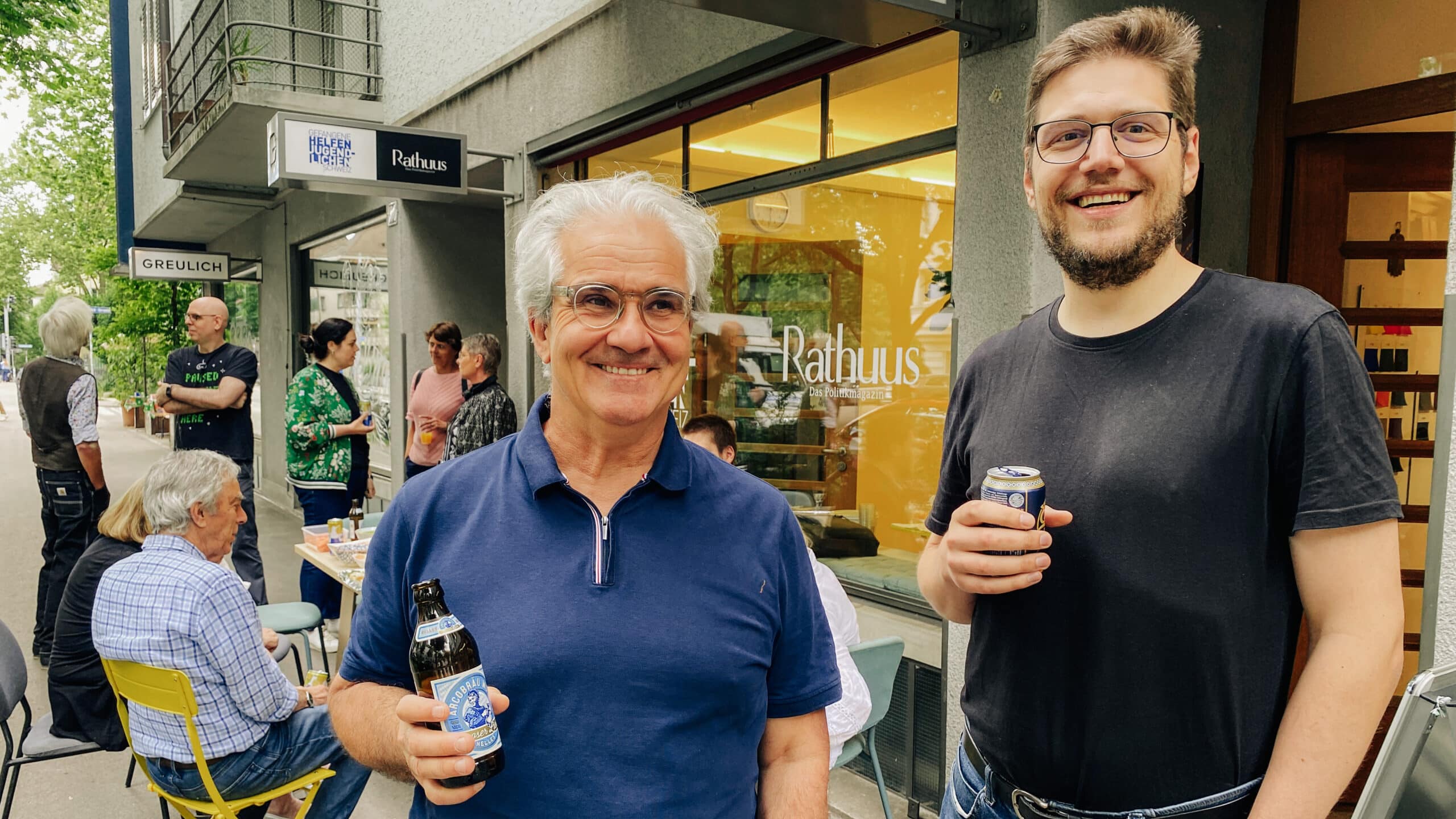 Lorenz Steinmann und Pascal Turin stehen vor ihrem Büro an der Stauffacherstrasse 180. Sie halten je ein Getränk in der Hand. Im Hintergrund sind die Gäste des Rathuus-Fäschtlis 2025 zu sehen.