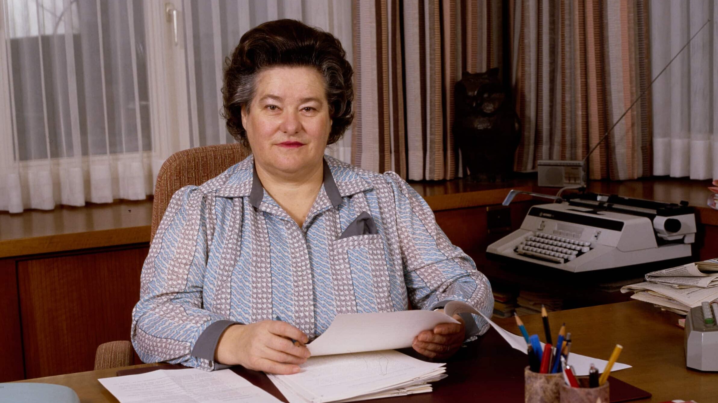 Hedi Lang sitzt 1987 an ihrem Schreibtisch. Vor ihr liegen Papiere. Neben ihr ist eine Schreibmaschine zu sehen.