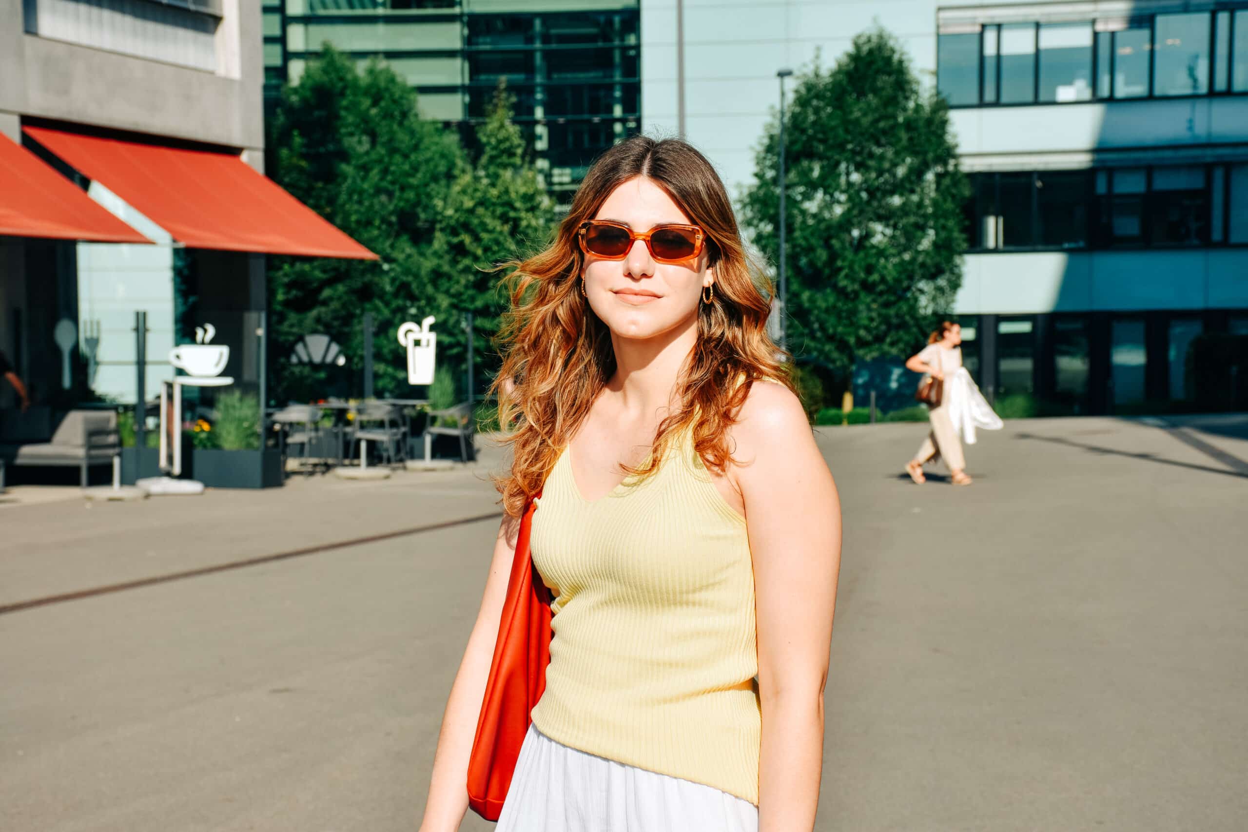 Leandra Columberg posiert für ein Foto in Stettbach. Es ist ein sehr sonniger Tag, darum trägt sie eine Sonnenbrille. Im Hintergrund sind ein Café, ein asphaltierter Platz und moderne Gebäude zu sehen.