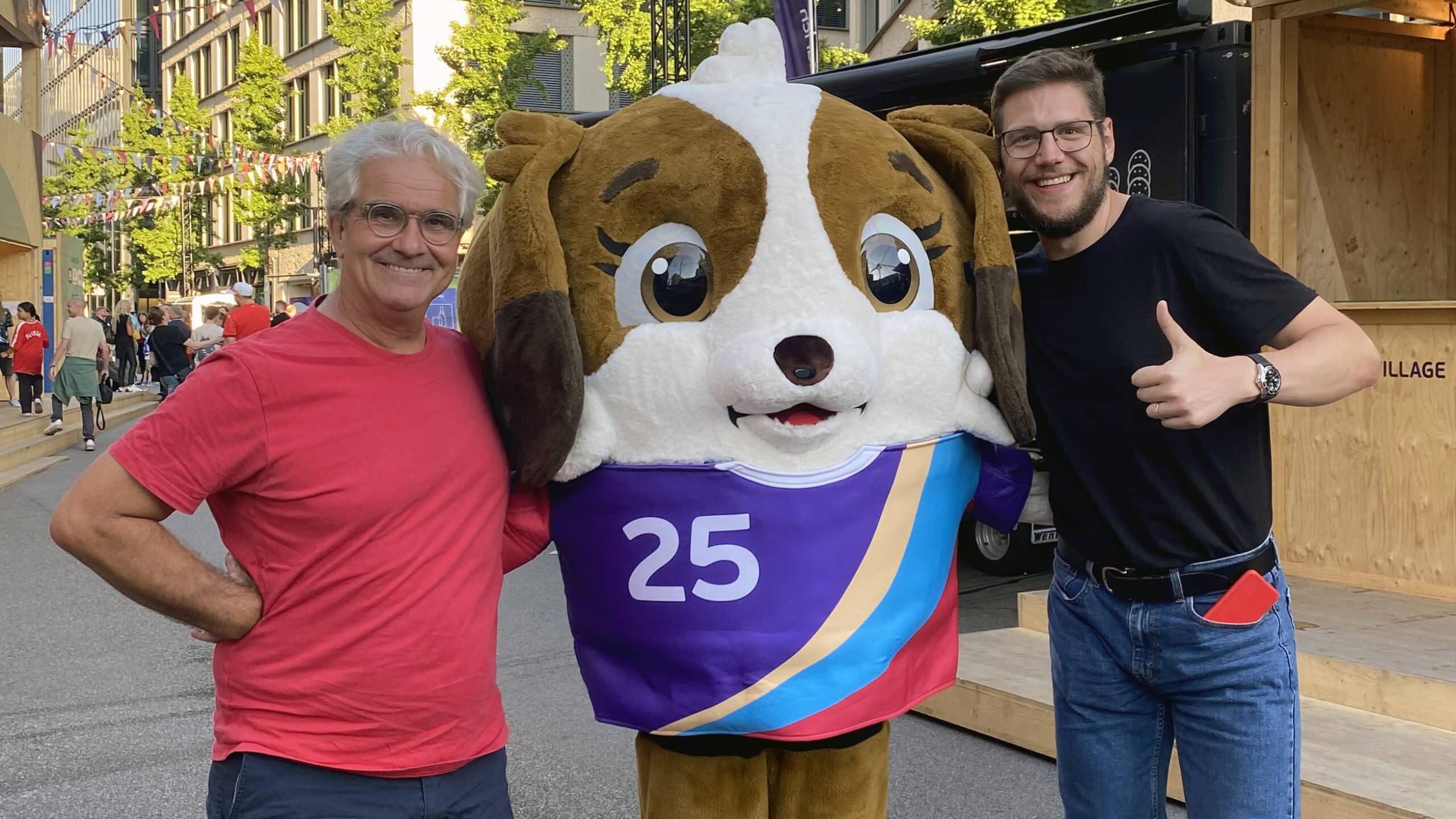 Lorenz Steinmann und Pascal Turin posieren locker, sommerlich gekleidet mit dem Maskottchen der Uefa Women's Euro 2025. Das Maskottchen soll ein Bernhardiner-Welpe darstellen.