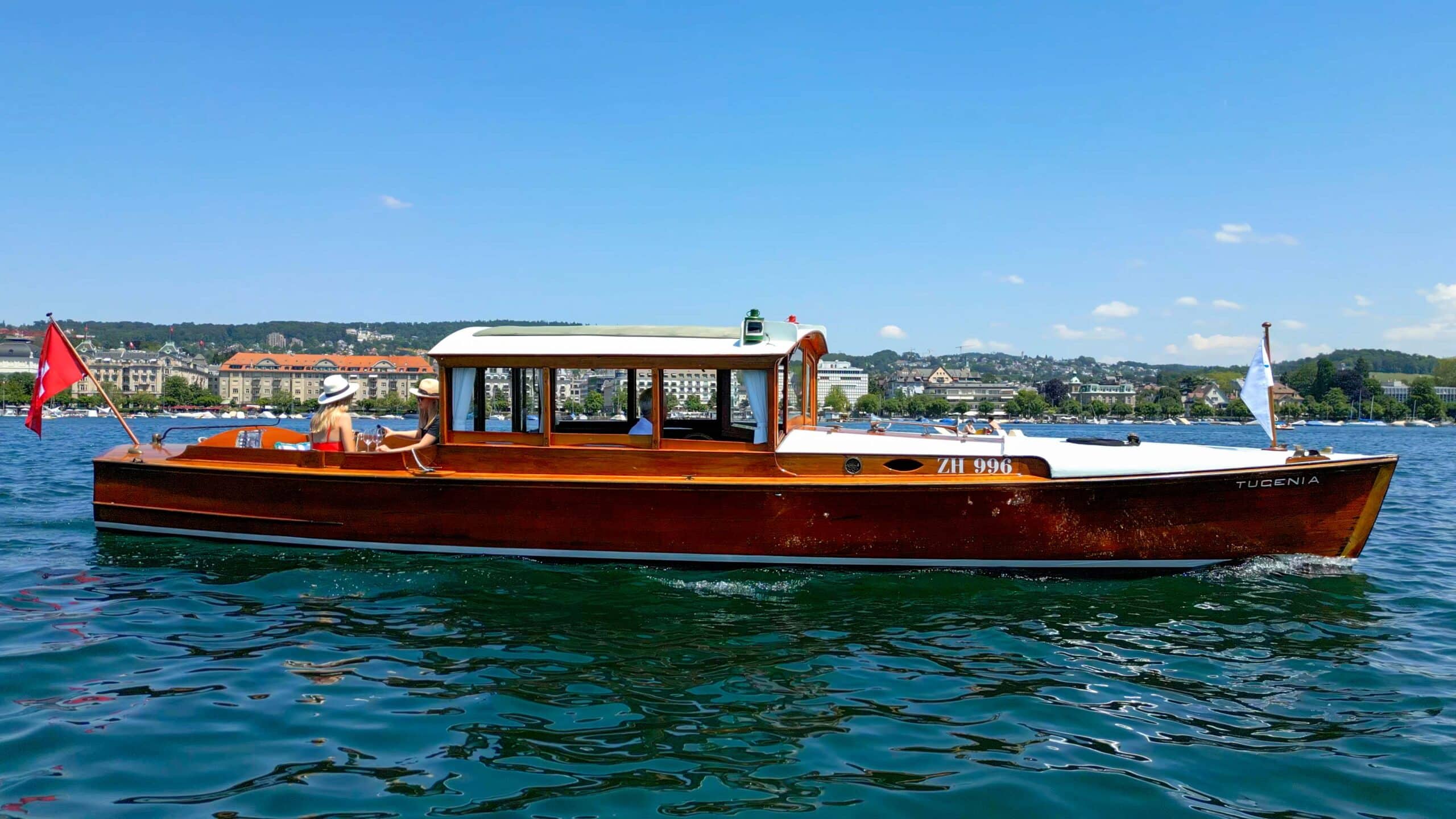 Das Elektro-Salonboot Tugenia fährt bei schönstem Wetter durchs Zürcher Seebecken. Zwei weibliche Gäste mit sommerlicher Bekleidung und Hüten sitzen hinten und geniessen die Fahrt.