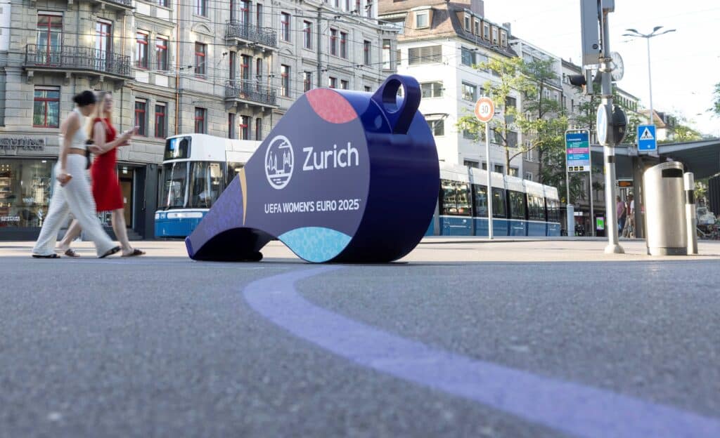 Eine überdimensionierte Trillerpfeife steht auf dem Löwenplatz. Zwei Frauen laufen vorbei, dahinter ist ein Tram der Linie 14 zu sehen.