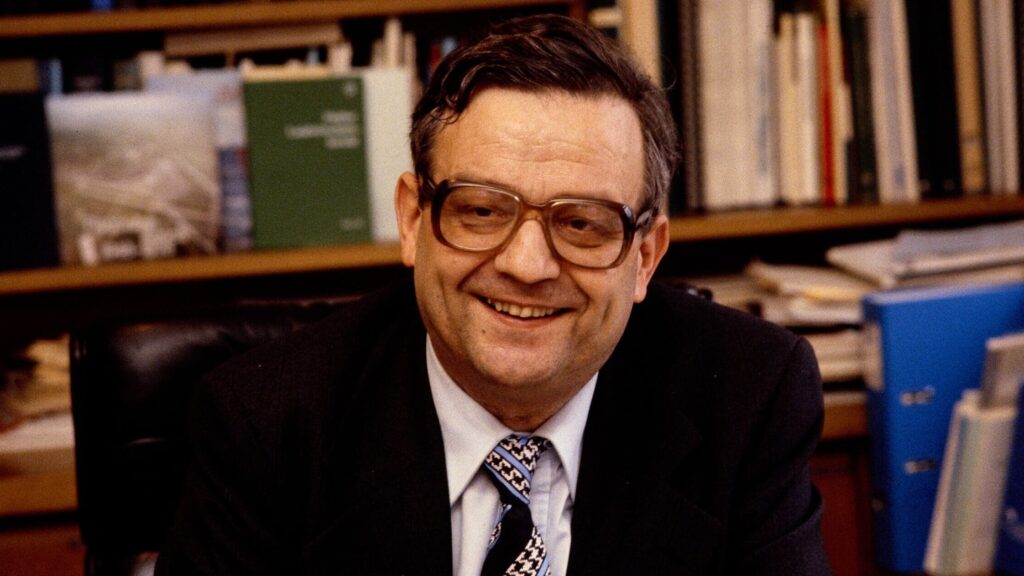 Hans Künzi sitzt 1971 mit Anzug und Krawatte in seinem Büro. Hinter ihm befindet sich ein Büchergestell voller Bücher. Hans Künzi trägt eine Brille und lächelt.