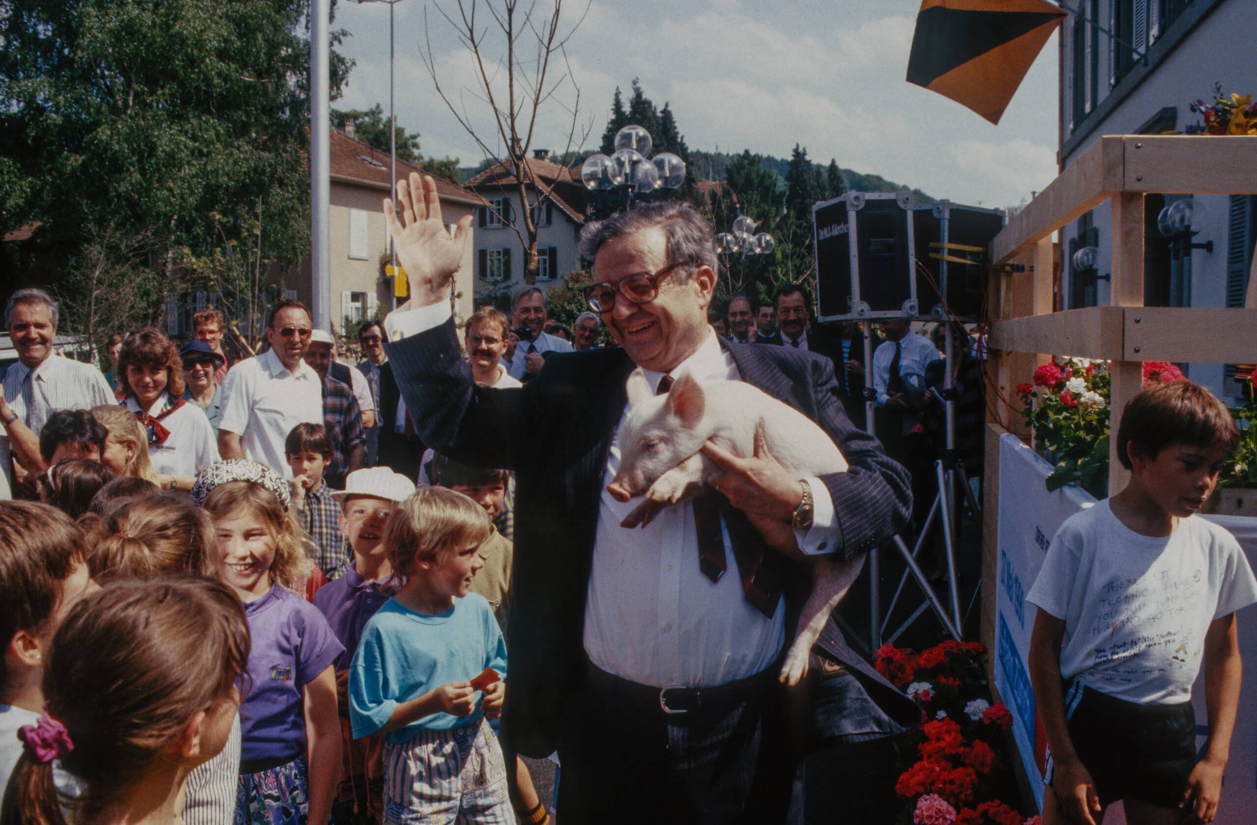 Hans Künzi im Anzug und Krawatte winkt der Bevölkerung an der Eröffnungsfeier der Zürcher S-Bahn zu. Im Arm hält er ein lebendes Schweinchen. Viele Kinder sind auf dem Bild zu sehen. Die Erwachsenen beobachten den Politiker. Hans Künzi wirkt glücklich.