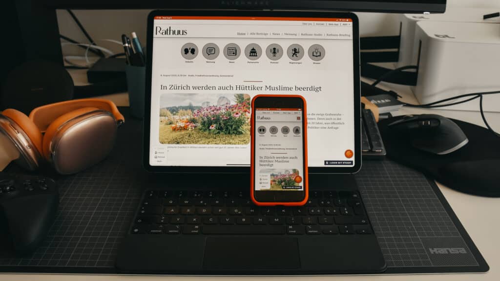 Ein iPad mit Tastatur und ein iPhone in roter Hülle mit auf den Bildschirmen eingeblendeter Web-App von Rathuus. Es handelt sich um eine Szene eines gut gefüllten Schreibtisches. Zu sehen sind eine Schreibmatte, Kabel, AirPods Pro, ein Xbox-Controller sowie eine Logitech-Maus. Die Geräte sind schwarz, die AirPods Pro orange. Die Schreibmatte ist ebenfalls schwarz, der Pult ist weiss.