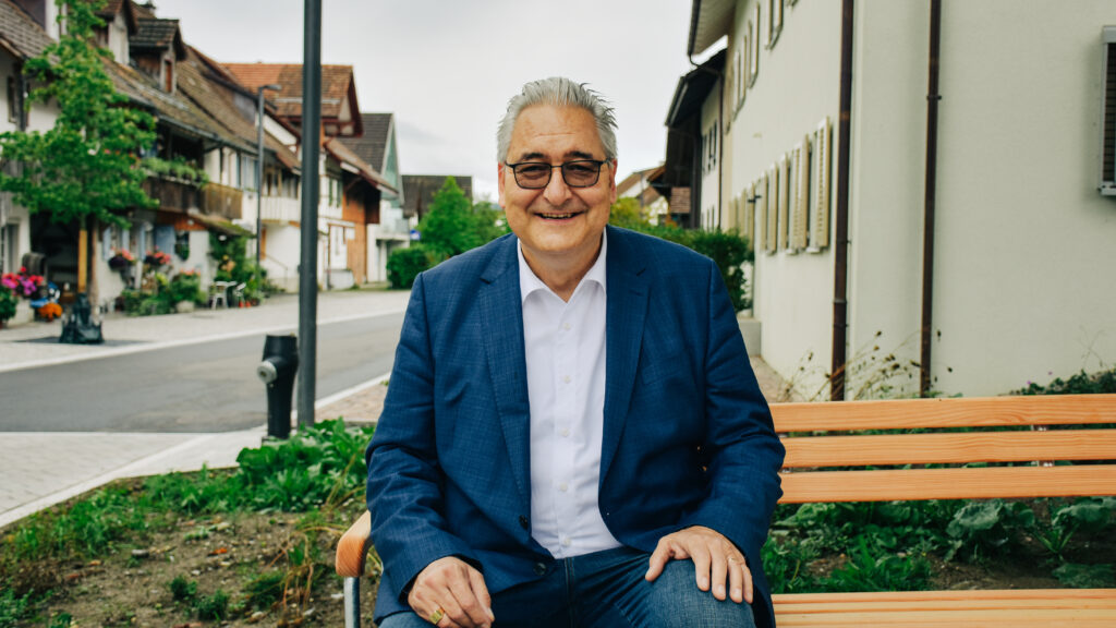 Beat Hauser, graue Haare und Brille, sitzt auf einer Parkbank im Rafzer Dorfkern. Hauser trägt ein blaues Jackett, ein hellblaues Hemd und Jeans.