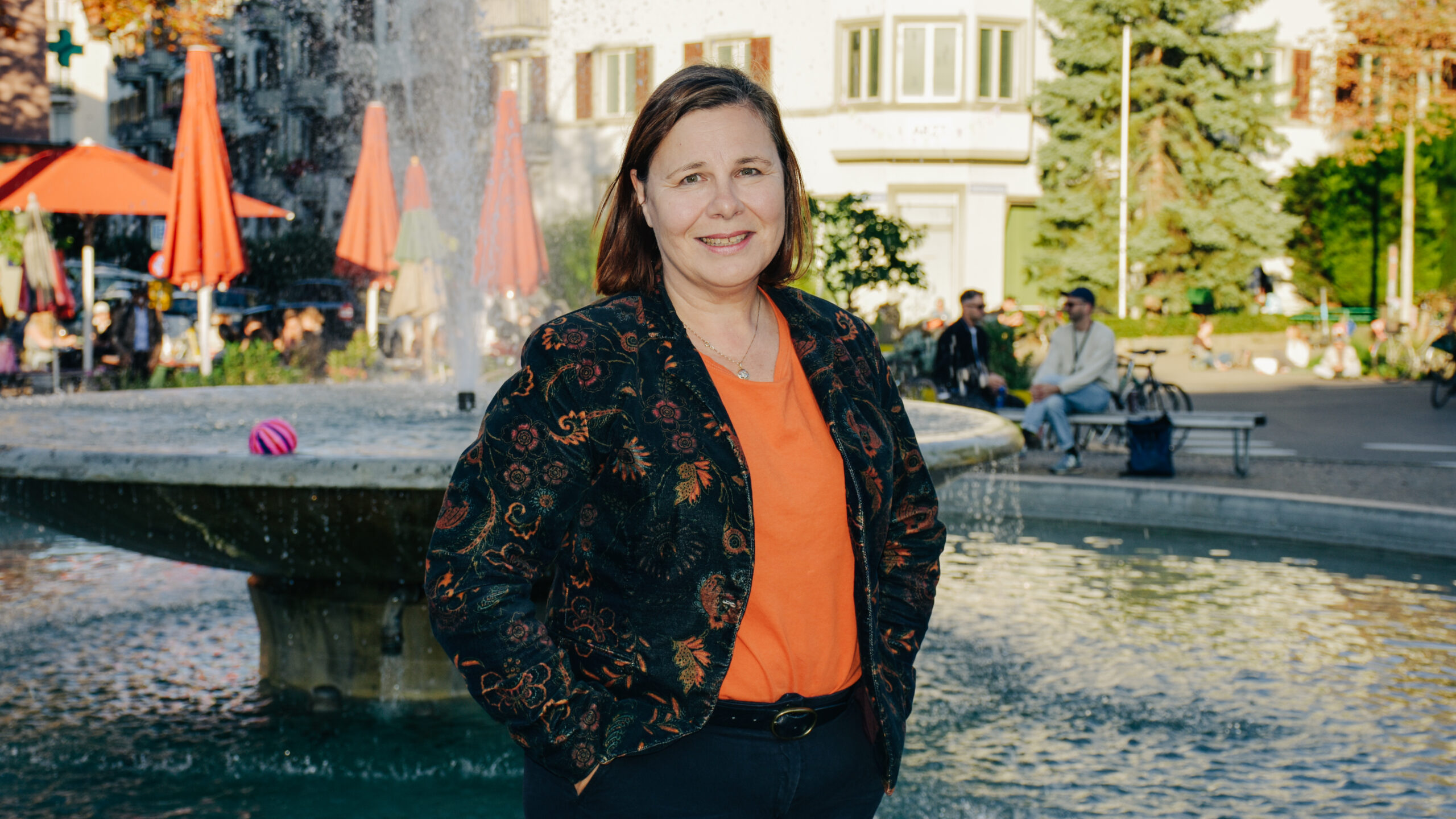 Kathrin Wydler steht vor dem Brunnen am Bullingerplatz. Sie trägt einen Blazer und ein oranges T-Shirt.