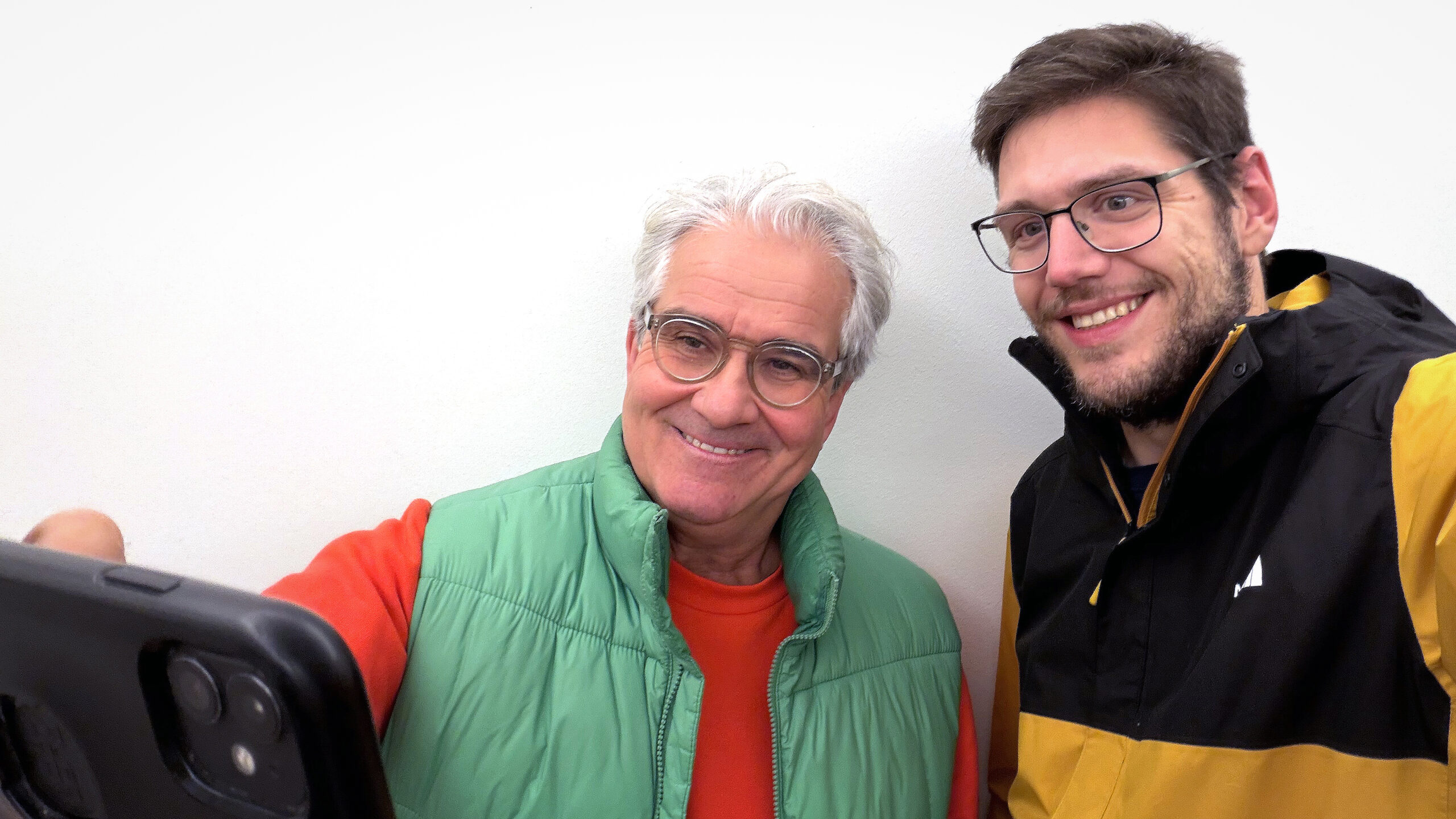 Lorenz und Pascal machen ein Selfie und schauen lächelnd in die Kamera. Lorenz trägt eine grüne Daunenweste, Pascal eine gelb-schwarze Übergangsjacke.