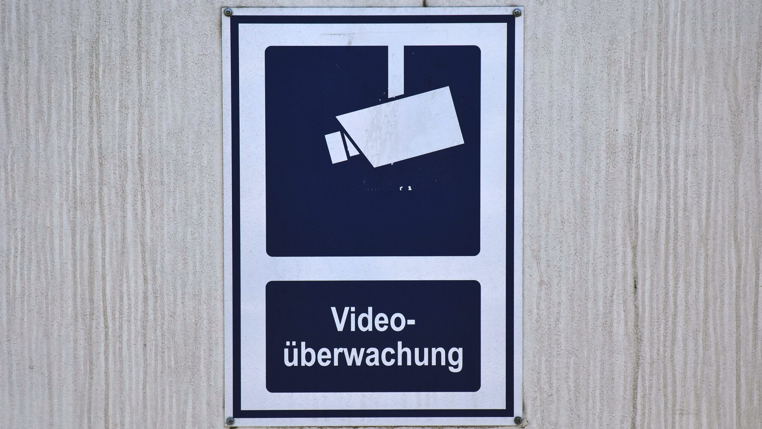 Ein dunkelblau-weisses Schild an einer Wand, dass eine Videokamera zeigt und den Text "Videoüberwachung".