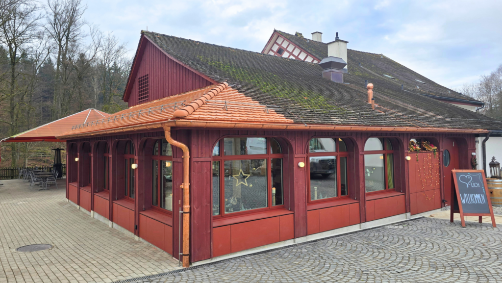 Rotes Restaurantgebäude, ein Willkommensschild steht vor der Eingangstür. Ein gepflasterter Bereich erstreckt sich bis zu einem Aussenbereich mit Sitzplätzen.