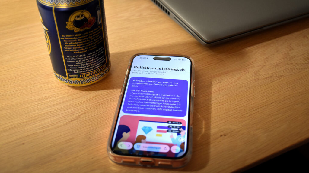 Ein Smartphone, das die Website Politikvermittlung.ch geöffnet hat, liegt auf einem Tisch aus Holz. Daneben steht eine Dose eines Softgetränks.
