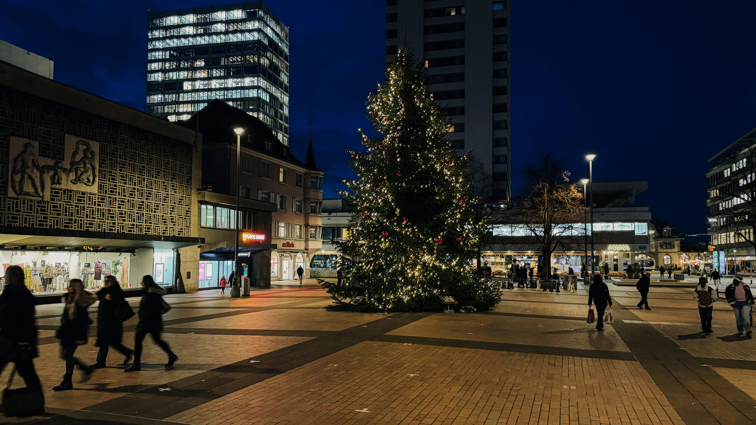 Ein mit Lichtern und Weihnachtskugeln behangener hoher Weihnachtsbaum steht auf dem Marktplatz Oerlikon. Es ist früher Abend und schon dunkel. Die Leute spazieren vom Shoppen vorbei. Im Hintergrund ragen zwei Hochhäuser empor.