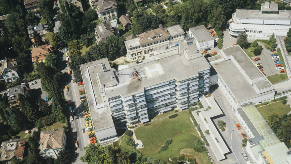 Das Kispi-Areal in Hottingen aus der Luft aufgenommen. Ein grosses Betongebäude ragt neben Einfamilienhäusern empor. Auf dem Dach steht ein Helikopter.