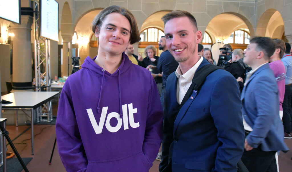 Zwei Vertreter der Kleinpartei Volt blicken lächelnd in die Kamera. Einer trägt einen violetten Pullover mit Volt-Logo, der andere einen blauen Anzug und ein weisses Hemd.