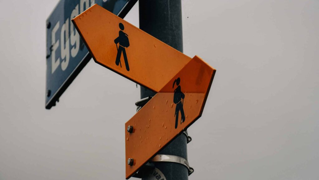 Zwei gelbe Wanderwegweiser, Regentropfen perlen an ihnen ab. Der Himmel ist grau.