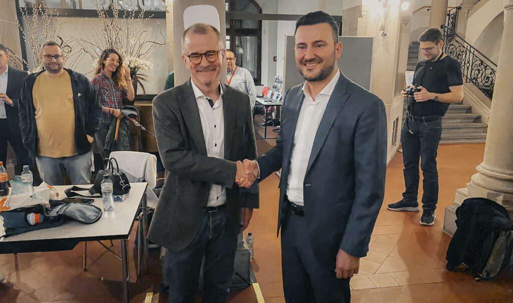 Zwei Männer mit Jackett schütteln sich die Hand und lächeln in die Kamera.