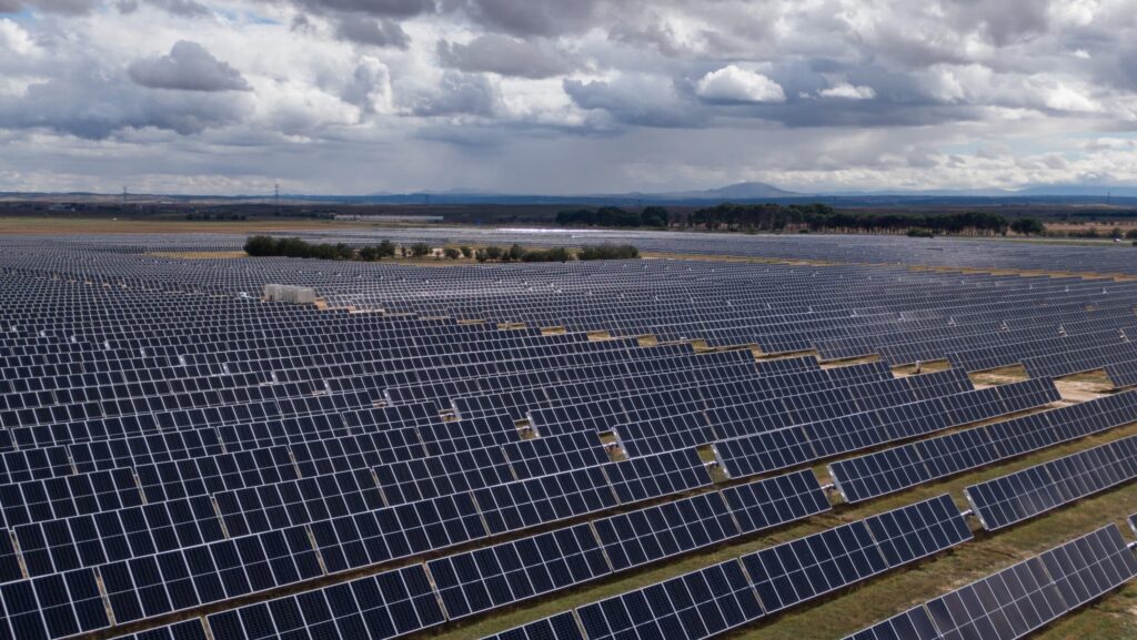 Eine grosse Solaranlage mit langen Reihen dunkler Solarpanels erstreckt sich über eine weite Landschaft unter einem bewölkten Himmel, mit Hügeln in der Ferne.