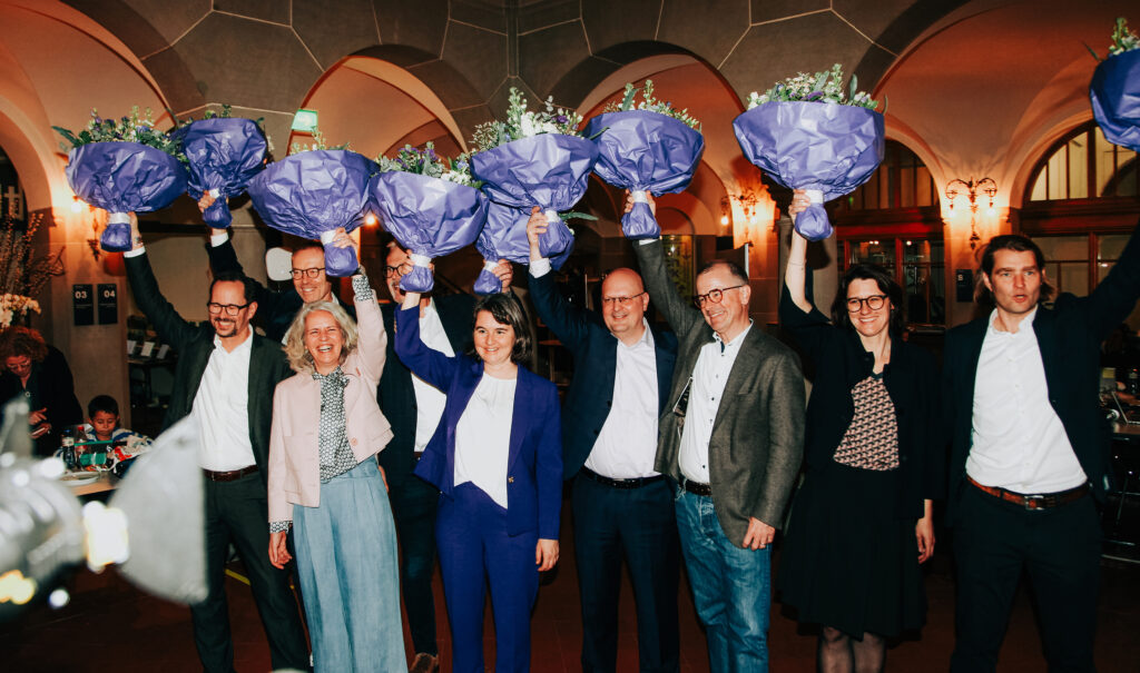 Die Gruppe frisch gewählter Stadträtinnen und Stadträte hält jubelnd Blumensträusse in die Höhe.