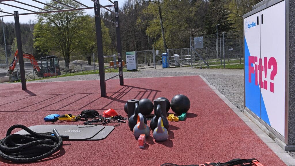 Outdoor-Fitnessgeräte liegen auf einer roten Tartan-Oberfläche vor einem Kasten mit der Aufschrift "Fit!?".