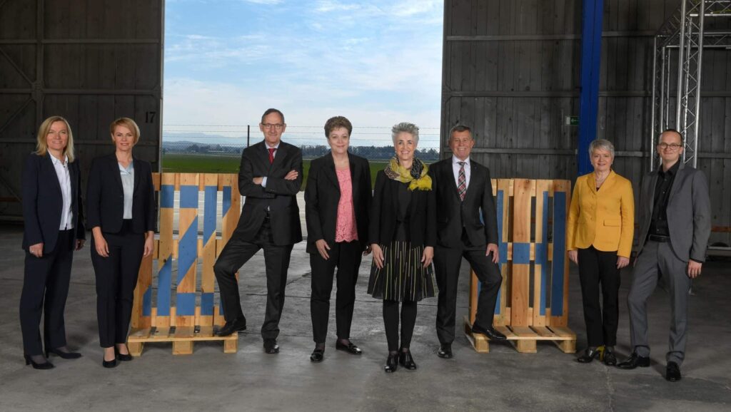 Zürcher Regierungsrat in einer offenen Halle mit Blick auf Landschaft. Die sieben Mitglieder posieren verteilt zwischen Holzpaletten.
