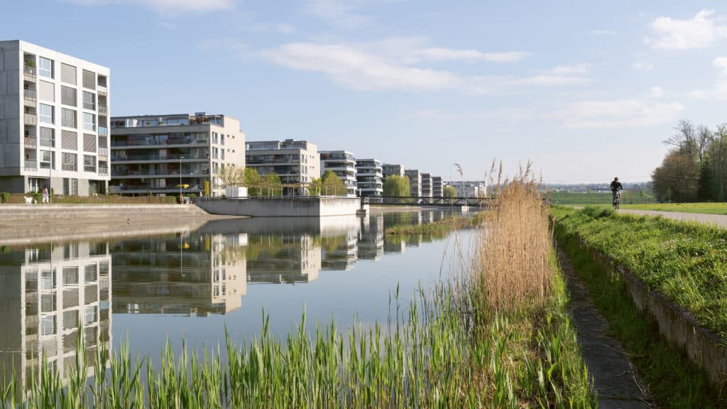 Moderne Wohnhäuser spiegeln sich in einem ruhigen Gewässer neben einem begrünten Weg.