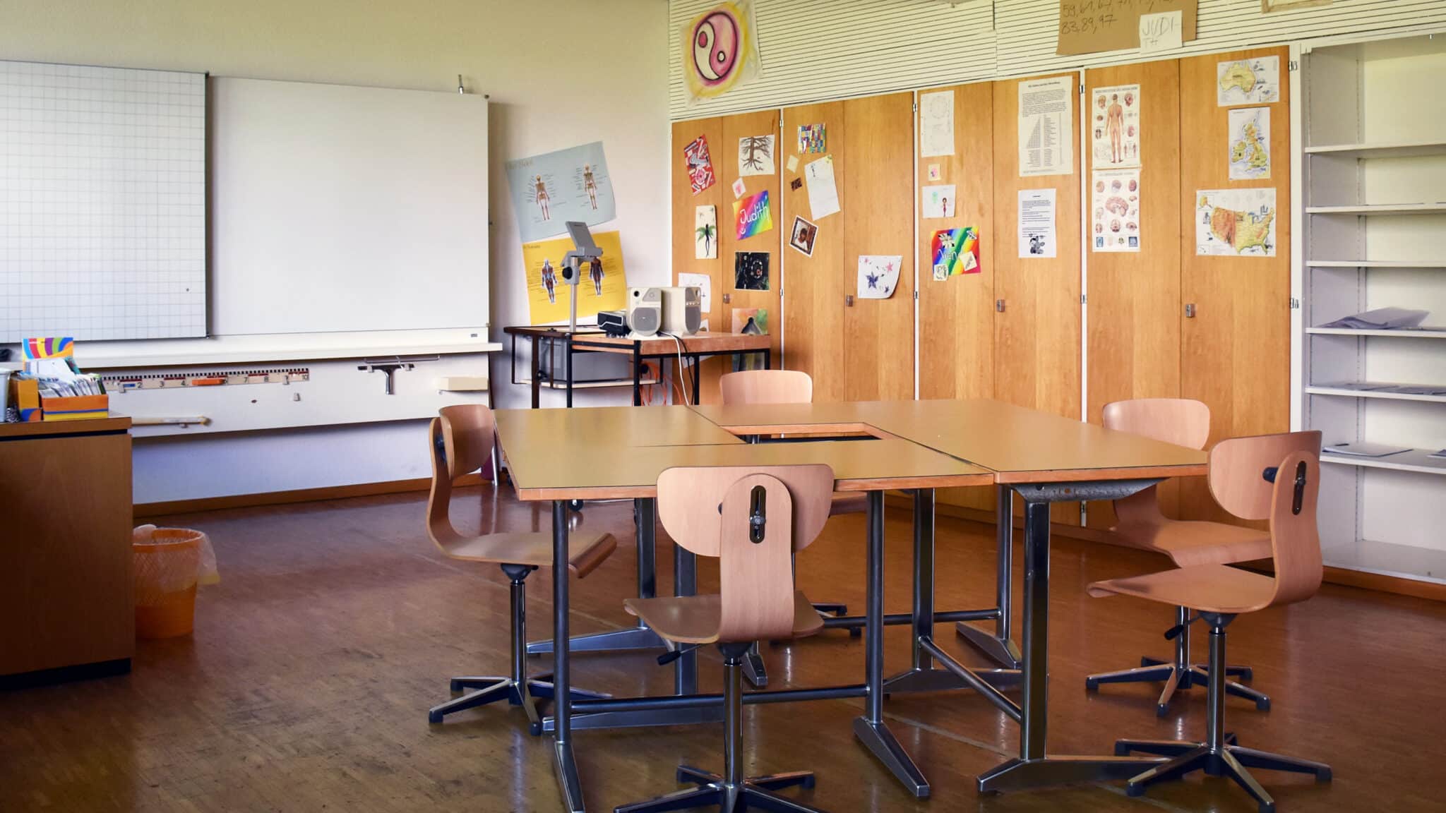 Leeres Klassenzimmer mit zusammengestellten Tischen, Stühlen, Wandtafel, Schulmaterial und Schränken mit Zeichnungen und Lernplakaten.