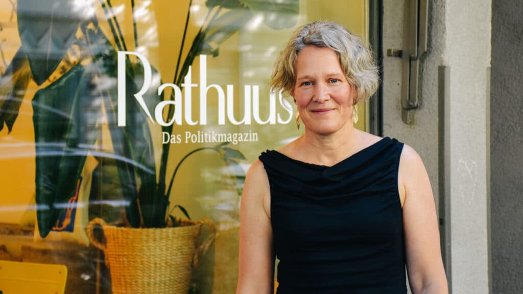 Frau mit kurzen grauen Locken in schwarzem Oberteil vor einem Schaufenster mit Pflanzen und dem Schriftzug „Rathuus – Das Politikmagazin“.