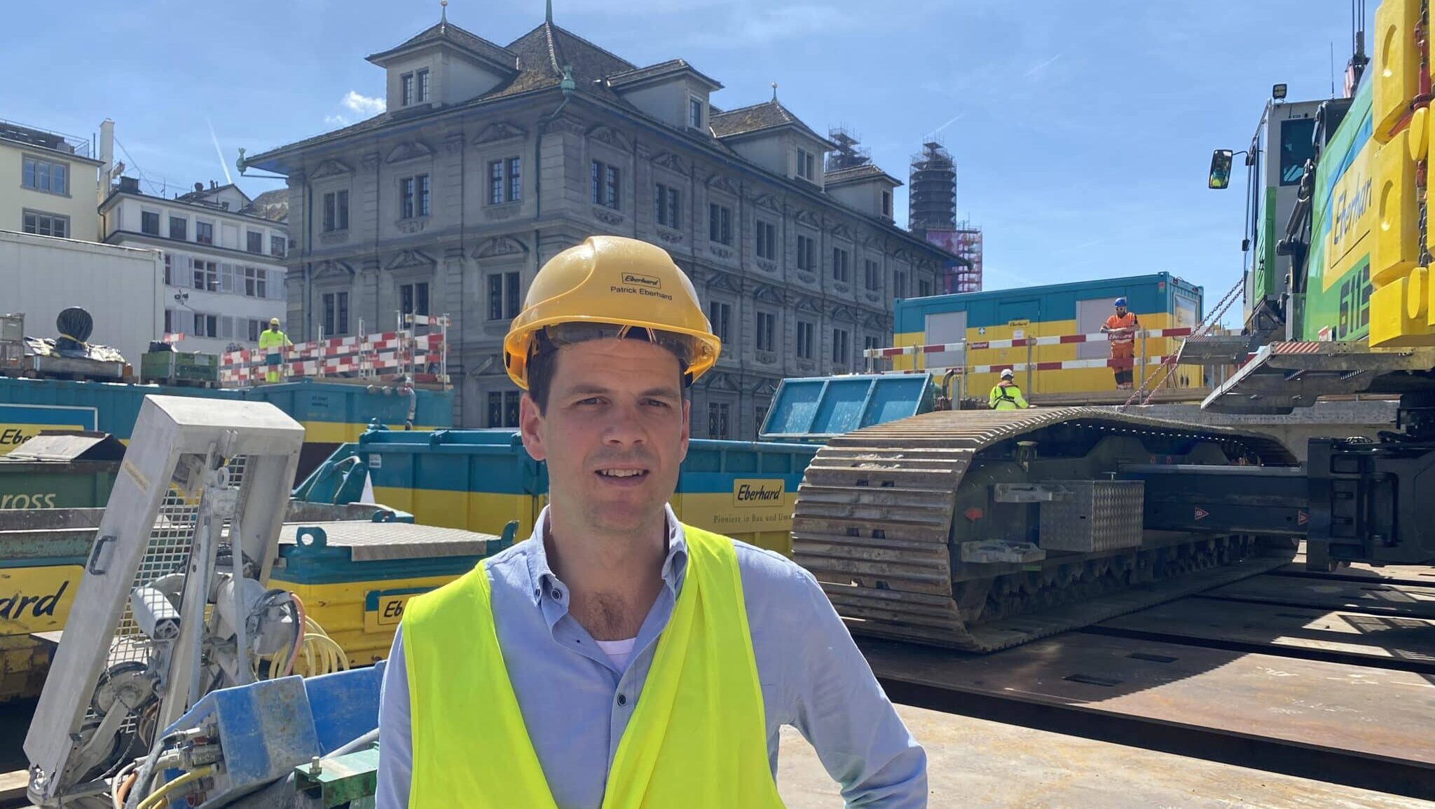 Zu sehen ist Patrick Eberhard, der CEO der gleichnamigen Baufirma. Er hat einen gelben Bauhelm auf und trägt eine gelbe Sicherheitsweste. Er steht auf der Rathausbrücke in Zürich, die aktuell umgebaut wird. Im Hintergrund zu sehen ist das historische Rathaus.