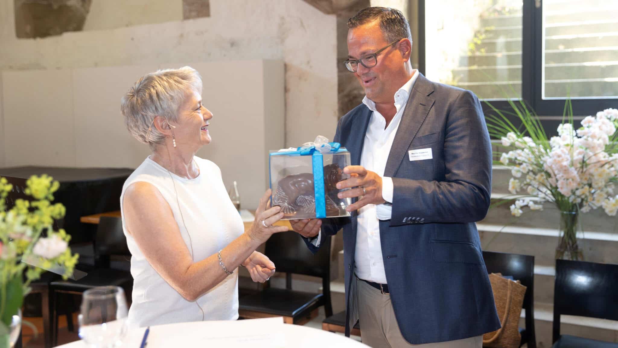 Eine Frau überreicht einem Mann bei einem Anlass ein verpacktes Geschenk mit blauem Band.