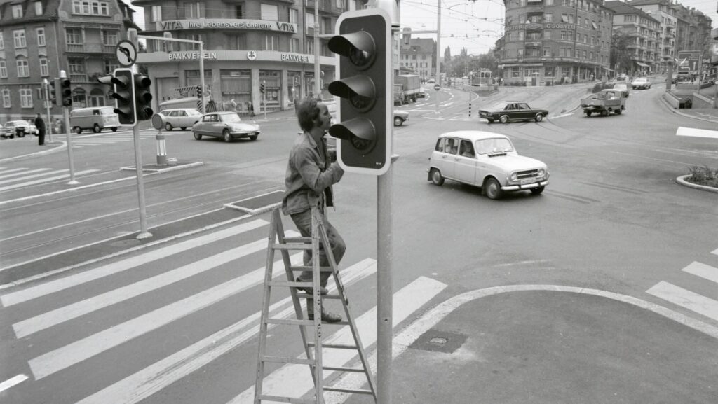 Zu sehen ist der Schaffhauserplatz auf einer Schwarz/Weiss-Aufnahme von 1971. Im Vordergrund eine Ampel, die gerade gewartet wird von einem Mann, der auf einer Leiter steht. über die Strasse tuckert ein Renault R4, im Hintergrund ist ein Citroén DS zu sehen.