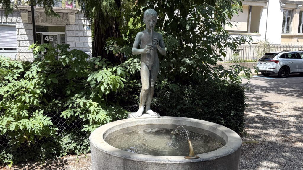 Zu sehen ist ein Brunnen in Zürich mit einem runden Becken und einem nackten Buben, der Flöte spielt. Gespiesen wird der Brunnen durch einen Wasserspender, an dem man auch trinken kann.