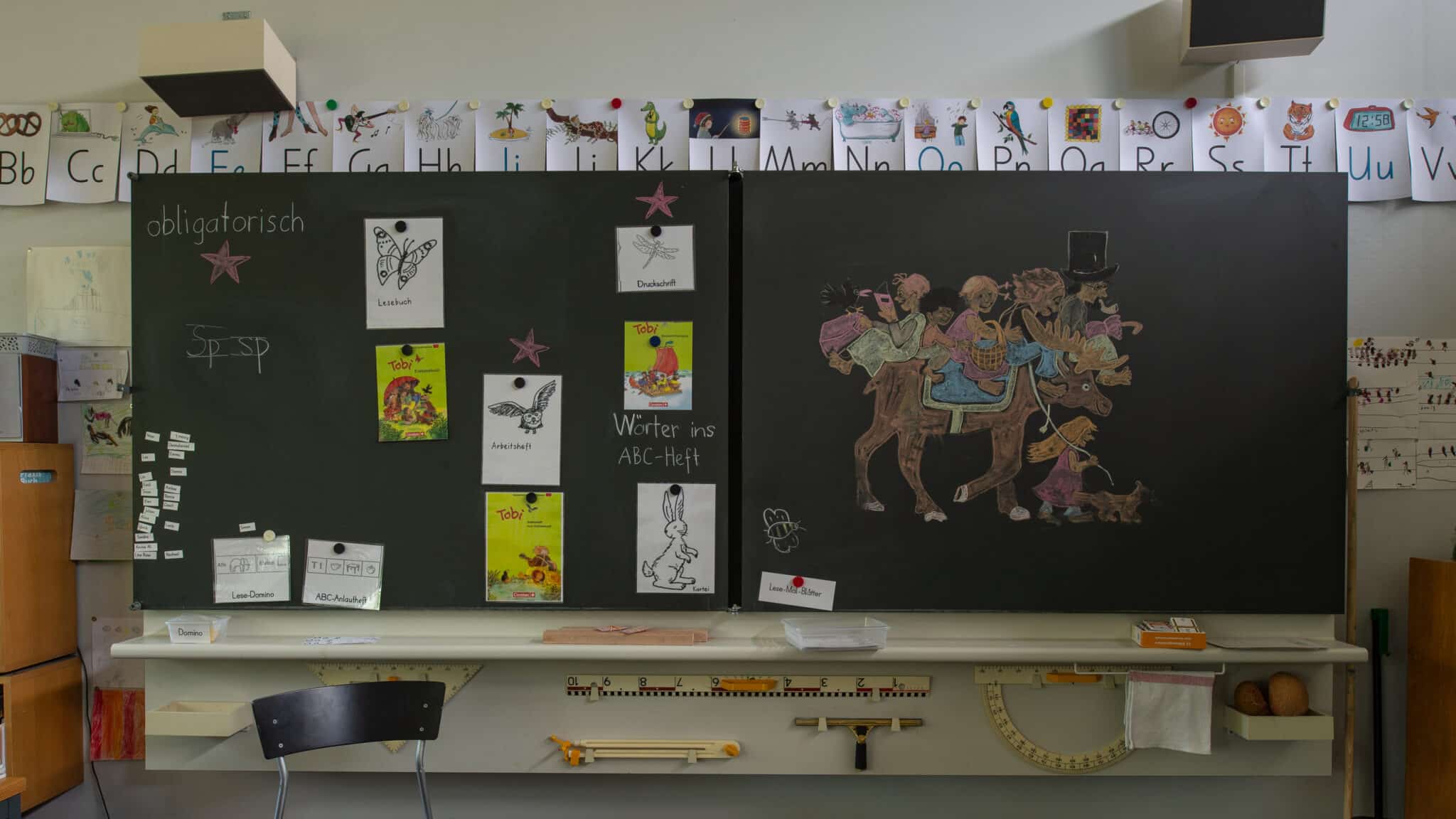 Klassenzimmer mit grosser Wandtafel, Lernmaterialien, Alphabetkarten und einer farbigen Kreidezeichnung.