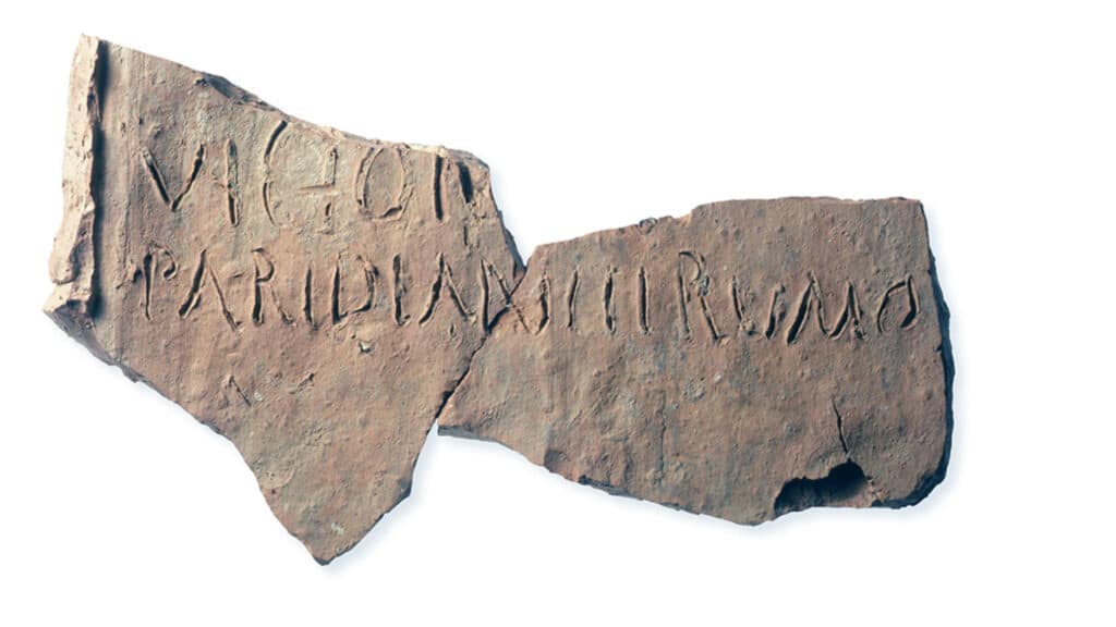 Fragment eines alten Leistenziegels mit eingeritzter lateinischer Inschrift auf hellem Hintergrund.