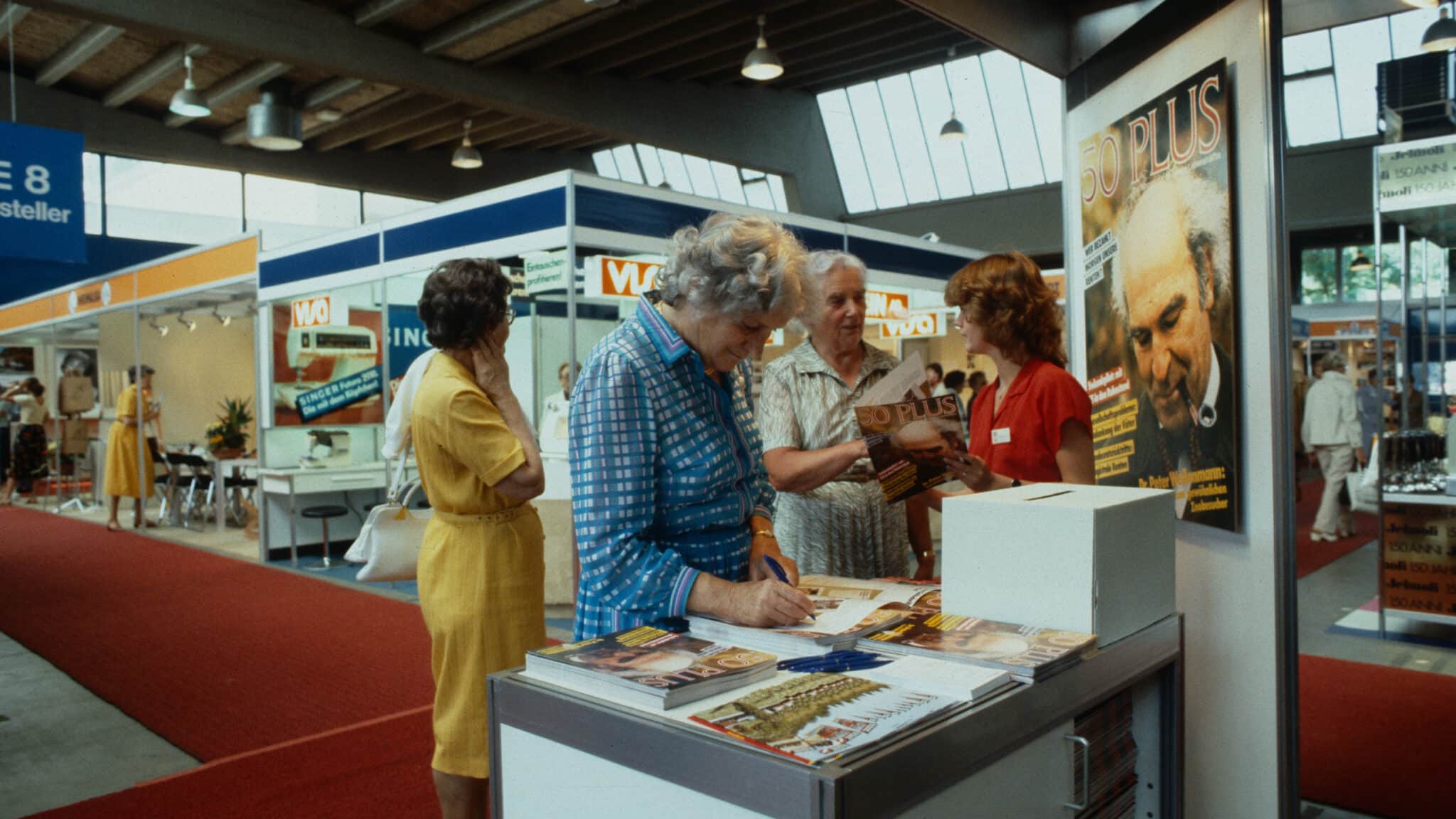 Altes Foto einer Seniorenausstellung mit Besucherinnen und Besuchern an einem Stand des Magazins „50plus“.