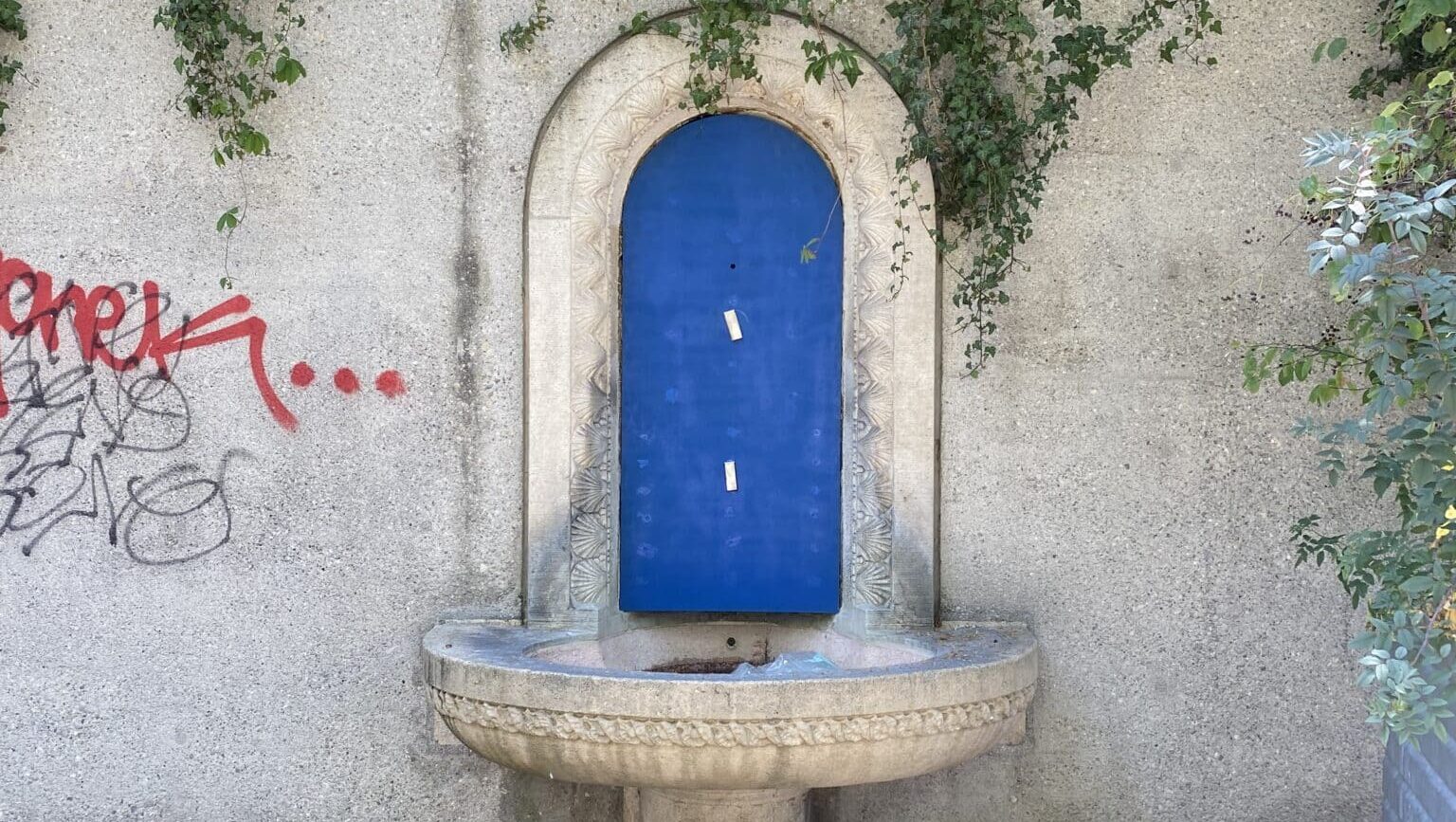 Zu sehen ist der Ort des Büebli-Brunnens an der Weinbergstrasse, wobei die Statue durch ein blaues Brett abgedeckt ist. Das Brunnenbecken aus Stein untendran ist leer.