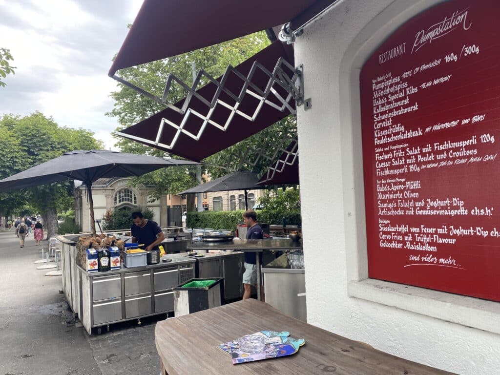Zu sehen ist die Beiz "Pumpstation" mit der Menuauswahl an der Wand im vordergrund und dem legendären Grill. Der Blick geht in Richtung Bellevue.