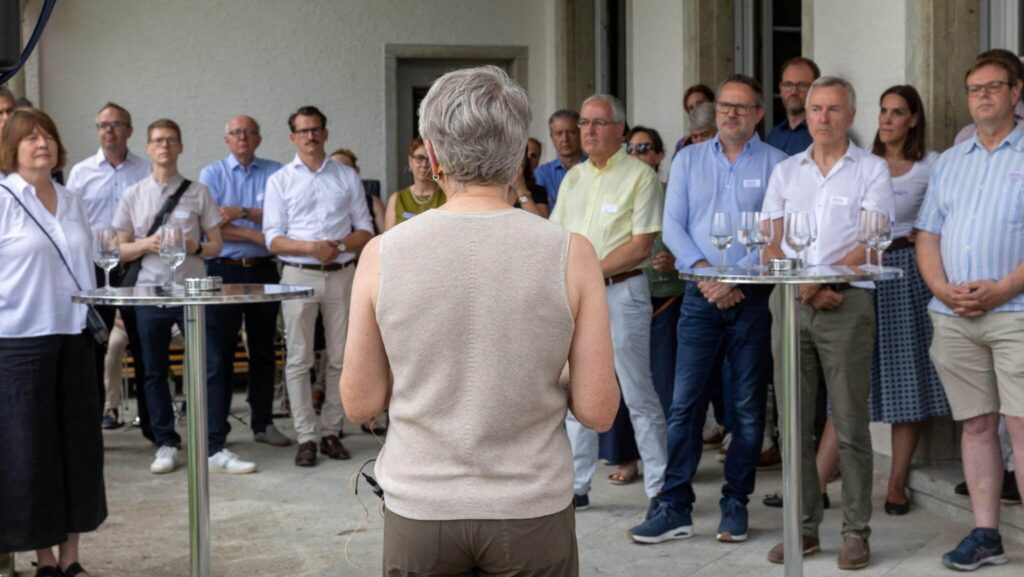 Frau spricht bei einem Anlass vor einer Gruppe von Zuhörenden auf einer überdachten Terrasse.