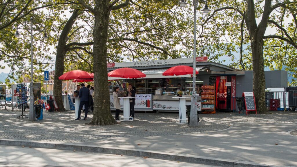 Zu sehen ist das "Bistro-Grill am See" mit drei roten Sonnenschimen im Vordergrund. Mächtige Bäume geben Schatten. Im Hintergrund verdeckt ist der Zürichsee zu erkennen.