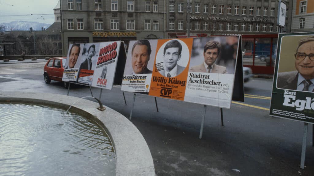Historische Wahlplakate mit Porträts von Kandidaten stehen an einer Strasse neben einem Brunnen.