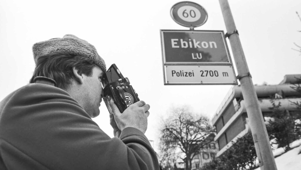 zu sehen ist ein Polizist in den 1980er Jahren, der ein Strassenschild mit einer Tempo-60-Tafel fotografiert. Auf dem Strassenschild steht Ebikon.