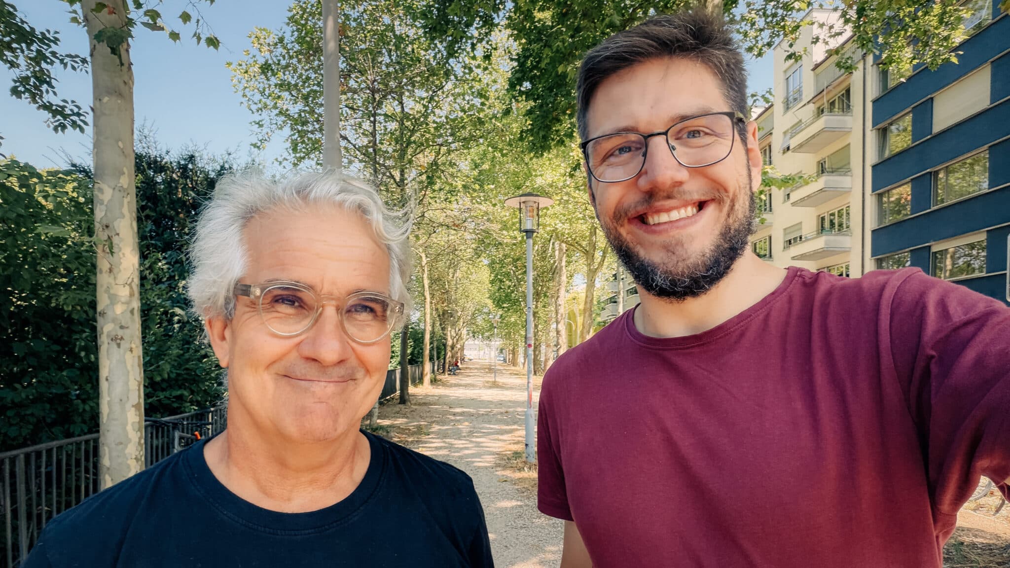 Zwei Männer machen ein Selfie auf einer sonnigen Strasse mit vielen Bäumen.
