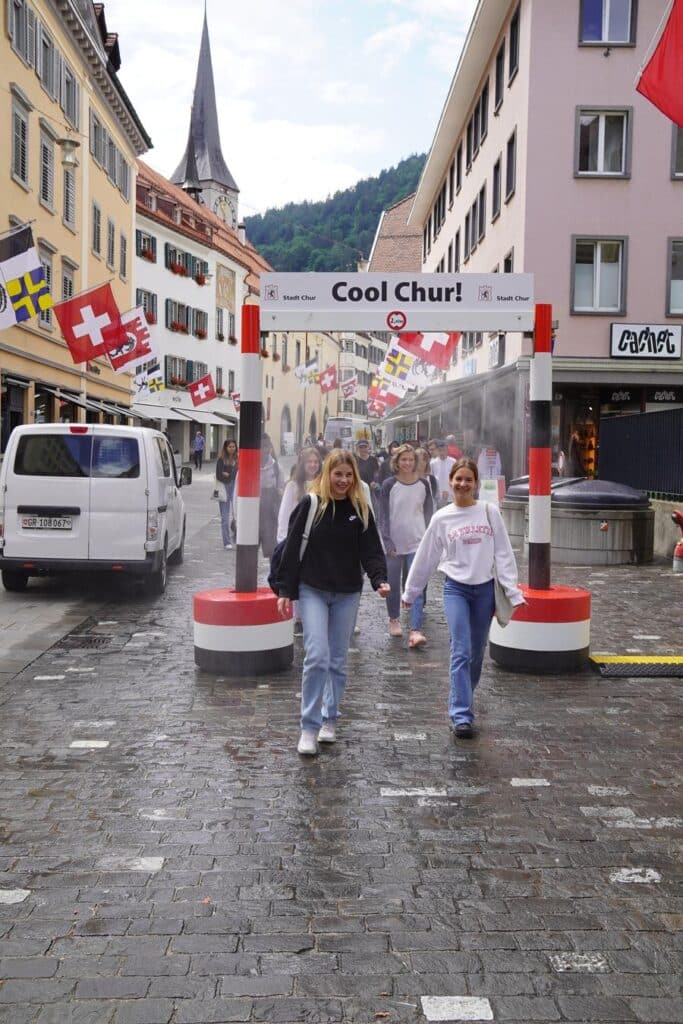 zu sehen ist eine Sprühdusche, die an zwei Betonpfeilern befestigt ist. Unten durch laufen einige junge Frauen, die sich sichtlich freuen über die Abkühlung.