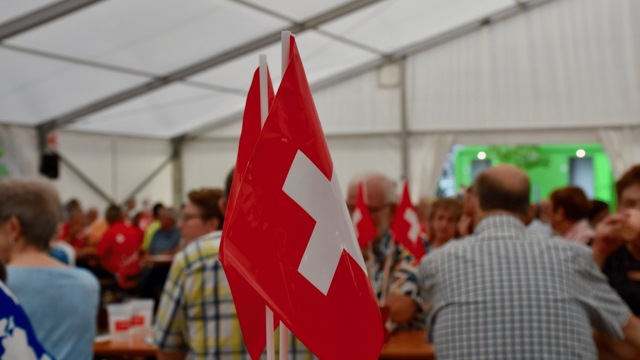 Zu sehen ist ein Symbolbild, eine 1.August-Feier in einem Zelt, diverse Leute von hinten fotografiert, im Vordergrund eine Schweizer Fahne.