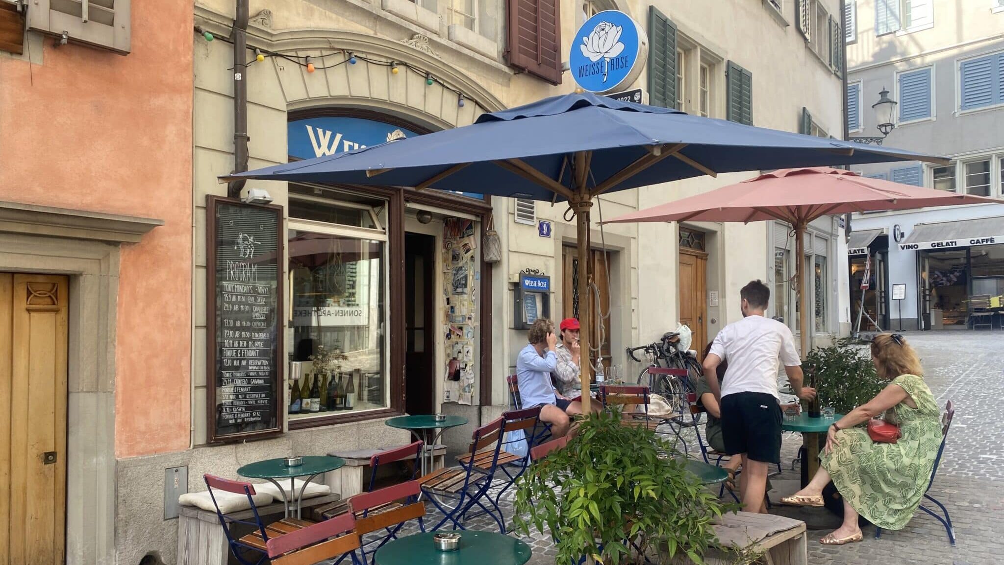 Aussenansicht des Restaurants "Weisse Rose" mit zwei grossen Sonnenschirmen, sowie mehreren Tischn und Bänken. Es hat einige Gäste und alles wirkt schön einladend.