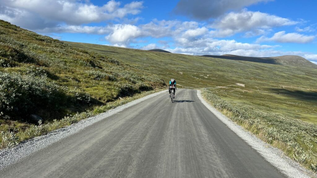 Die endlosen Weiten von Norwegen, mit Naturstrasse und einem Velofahrer, der aus der Ferne zu sehen ist. Die hügelige Landschaft ist grün mit ein wenig Buschwerk.