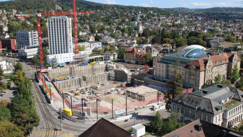 Von oben zu sehen ist die 30 Meter tiefe Baugrube, wo das Forum UZH gebaut wird. Rechts zu sehen sind drei rote Baukräne, im Hintergrund steh das Hochhaus des Instituts für medizinische Genetik. Das Wetter: schön!