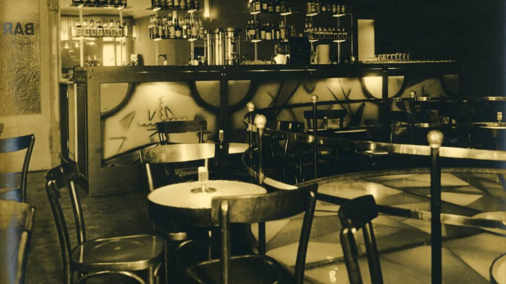 Zu sehen ist eine schwarz-weisse Aufnahme aus dem Innern des Maxim etwa im Jahr 1940. Es hat einige Tische und Stühle, hinten eine Bar, sowie rechts in der Bildmitte eine erhöhte runde Bühne mit Geländer.