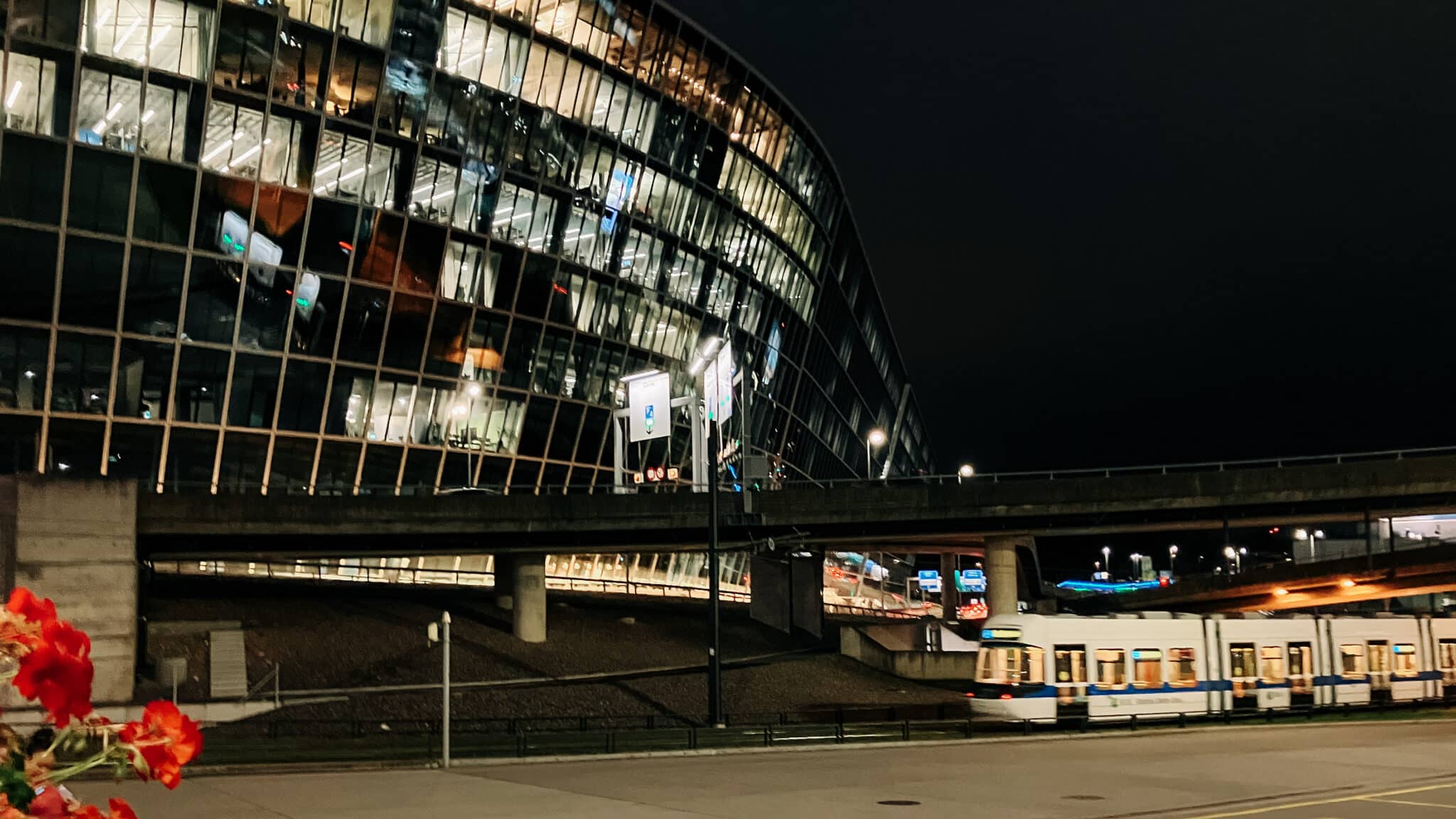 Beleuchtetes Gebäude am Flughafen Zürich bei Nacht, davor fährt ein Tram vorbei.